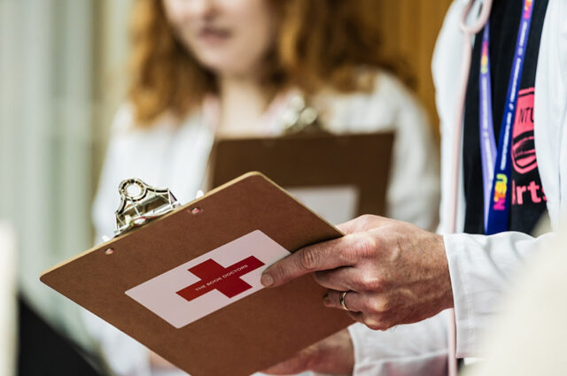 A hand holding a wooden clipboard with a white sticker and red cross on the back.